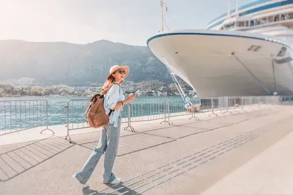 woman in sun hat and wearing a backpack, jeans and a shirt about to board a cruise ship on a sunny day